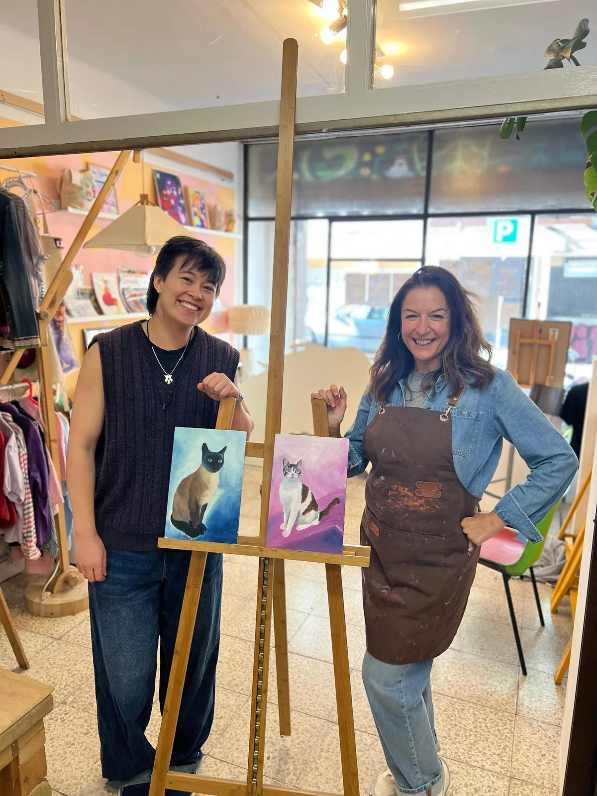 Two smiling people at the studio easel, each with a finished painting: a Siamese cat on a blue ground and a white and brown cat on a pink ground.