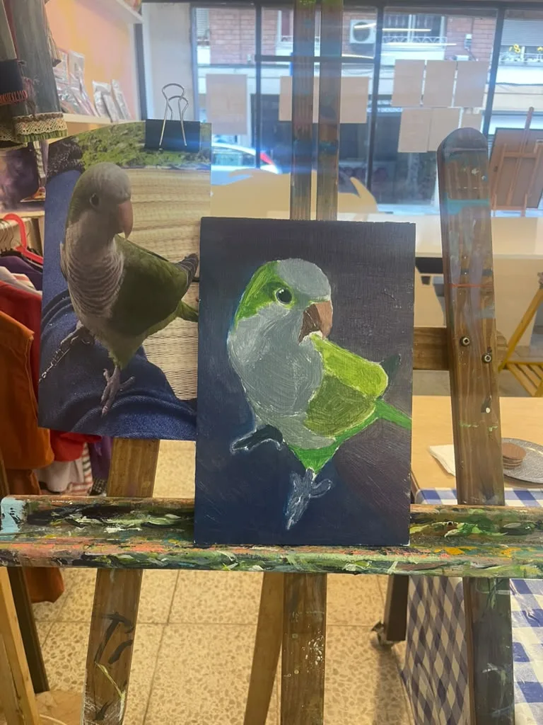 A finished painting of a green parakeet on a dark blue ground, beside the owner’s photo.