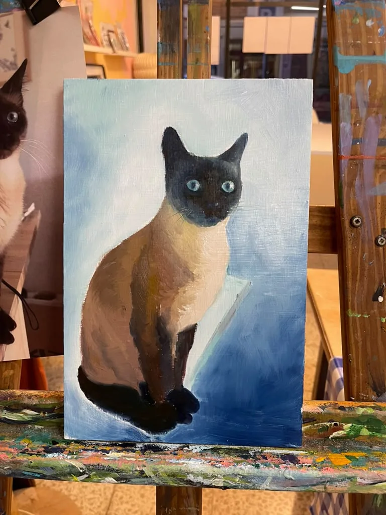 A finished oil painting of a Siamese cat on a pale blue ground, beside the owner’s photo.