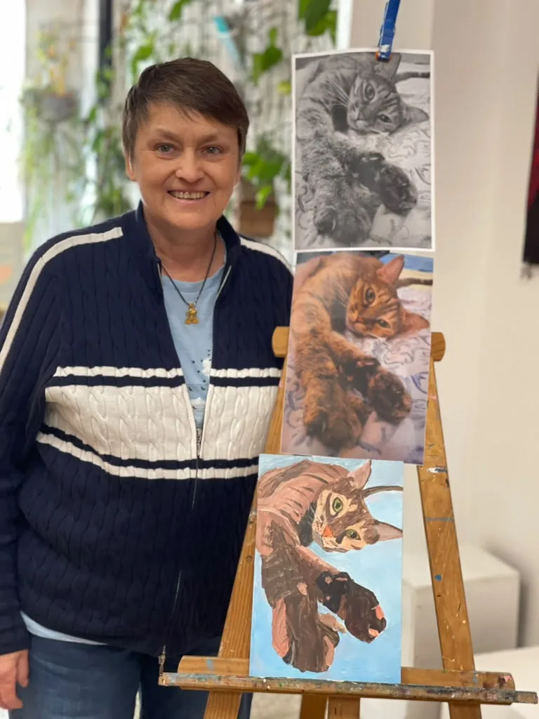 A smiling visitor beside the easel, with the reference photo of a tabby cat above the finished painting of the cat on a pale blue ground.