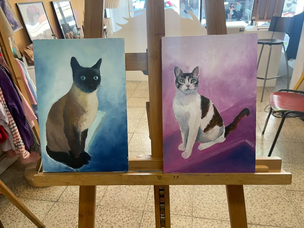 Two finished paintings side by side on the easel: a Siamese cat on a blue ground and a white and brown cat on a purple ground.